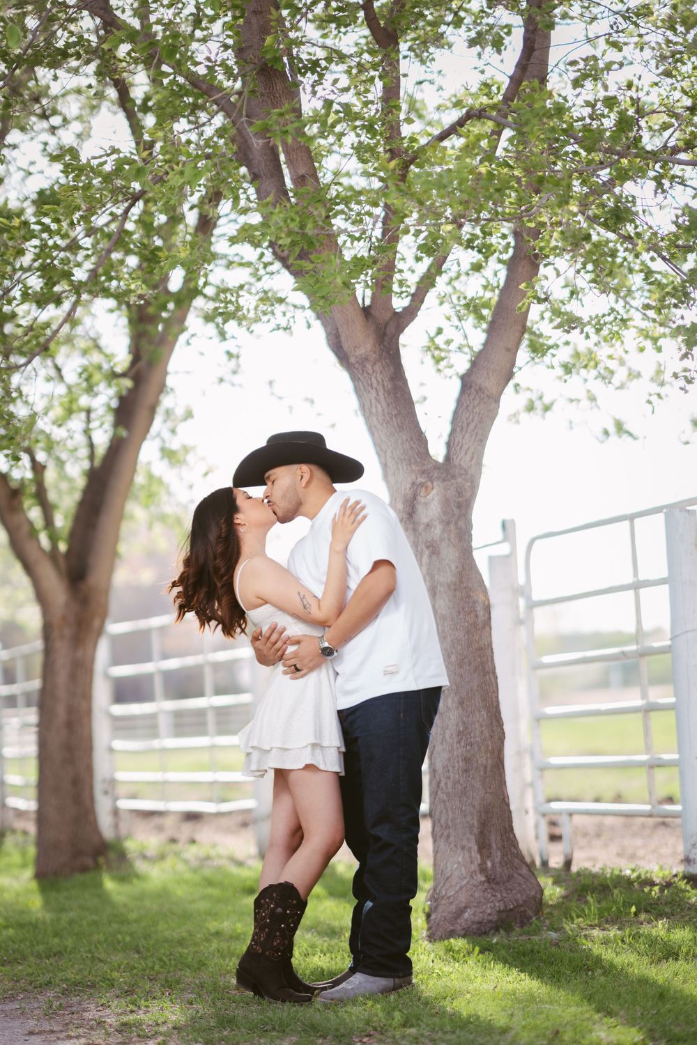Couple sharing a romantic kiss under green trees, woman in white dress and cowboy boots, man in black cowboy hat.