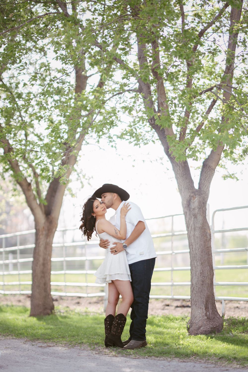 Couple embracing and kissing outdoors under green trees near a farm fence, woman in white dress and cowboy boots.
