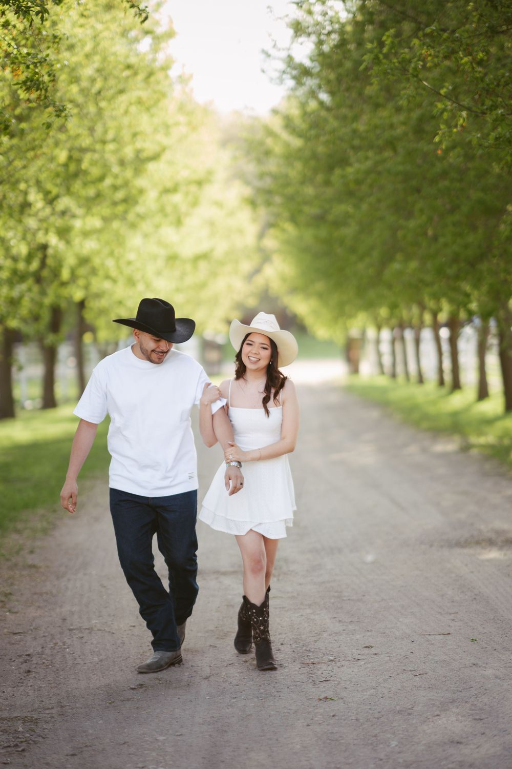 Couple in cowboy hats walking down a tree-lined dirt path, woman in white dress and boots smiling.