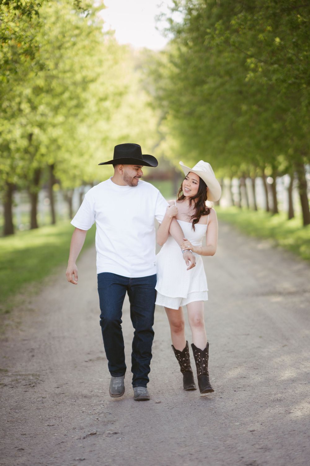 Couple in matching cowboy hats strolling a scenic tree-lined path, woman in white dress and cowboy boots.