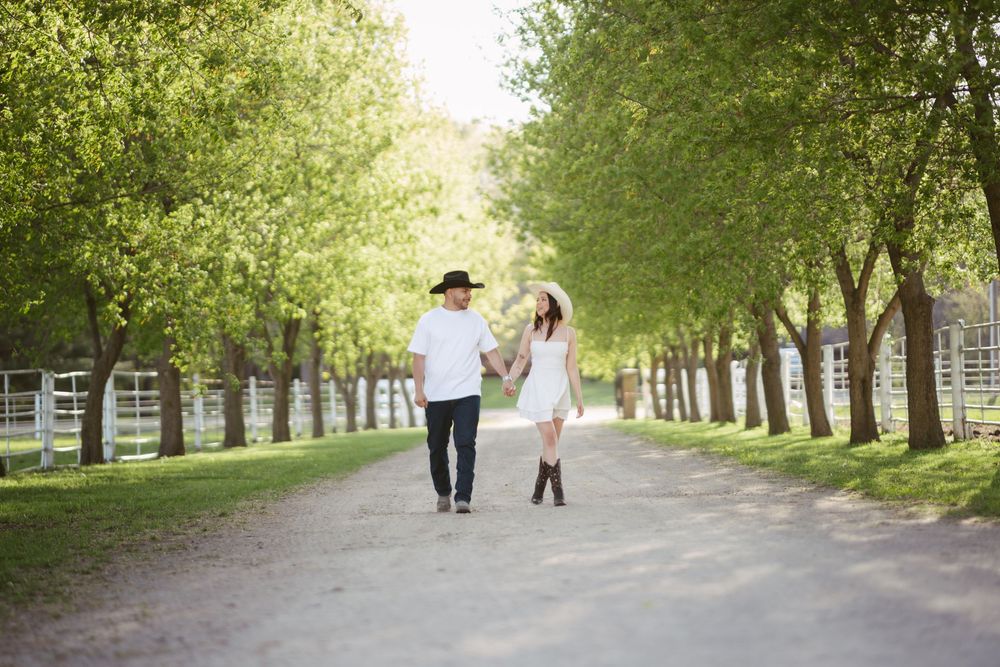 Couple holding hands walking down a tree-lined path in a sunny park, woman in white dress and man in cowboy hat.