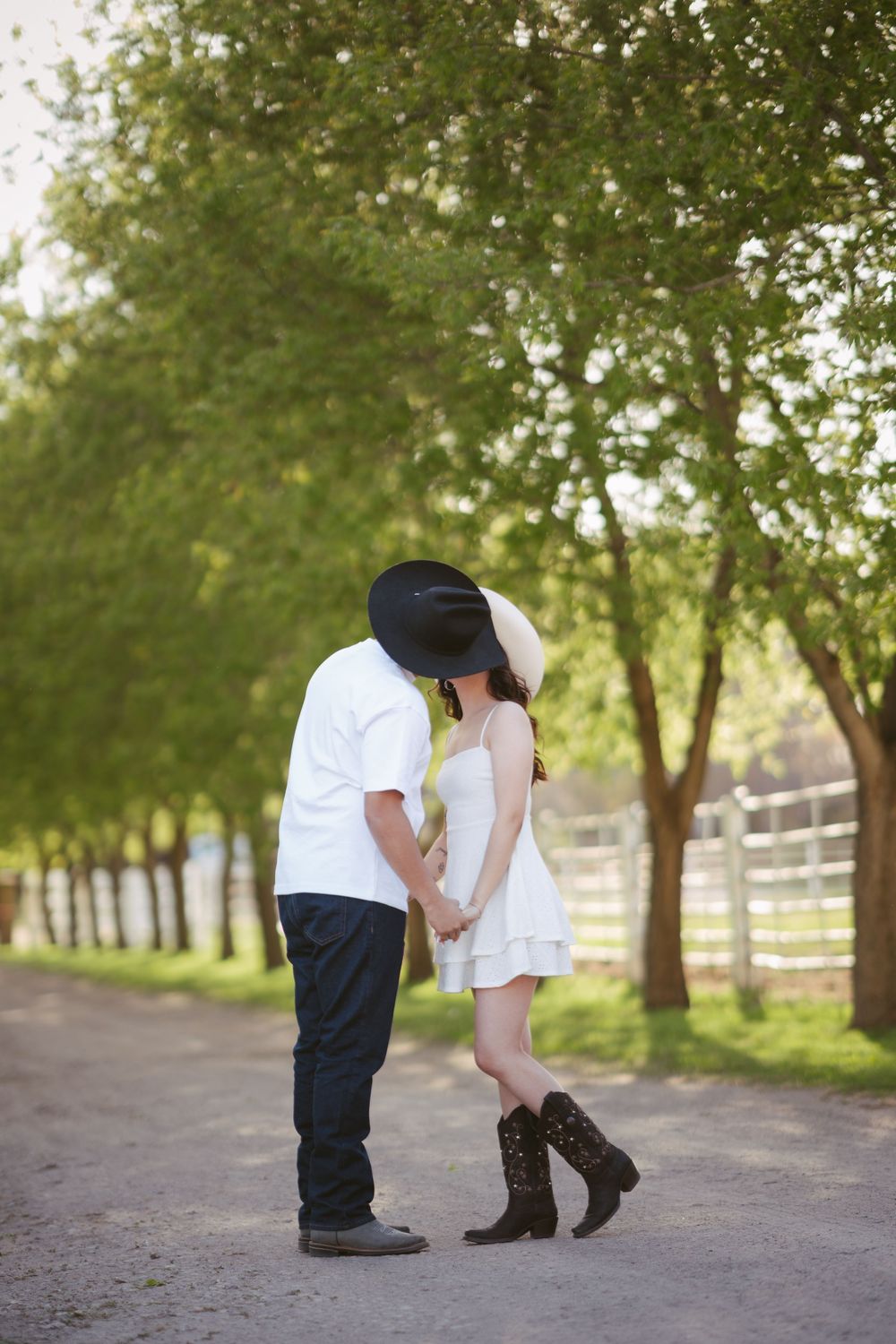 Couple kissing on a tree-lined path, wearing cowboy hats and boots, holding hands in a romantic outdoor setting.