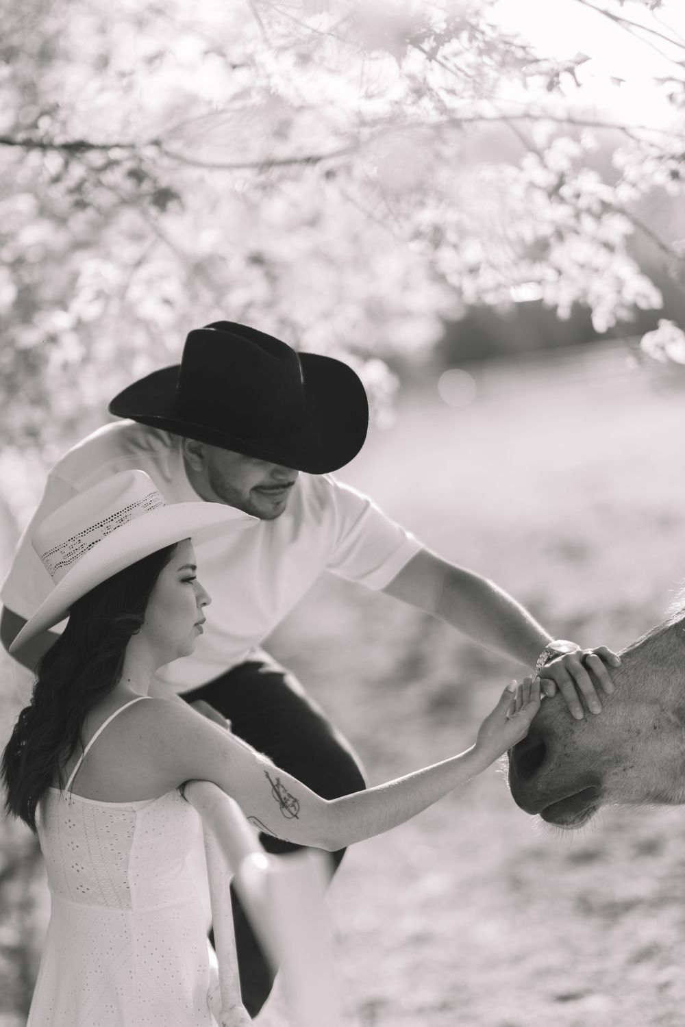 Black and white photo of couple in cowboy hats petting a horse under blossoming trees on a ranch.