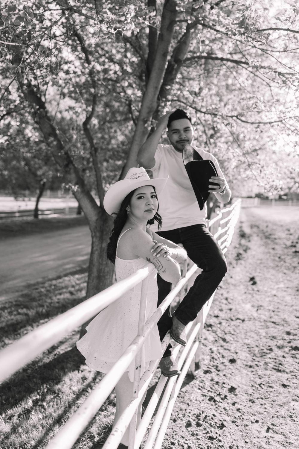 Black and white photo of a stylish couple posing by a white fence under blooming trees in a park.
