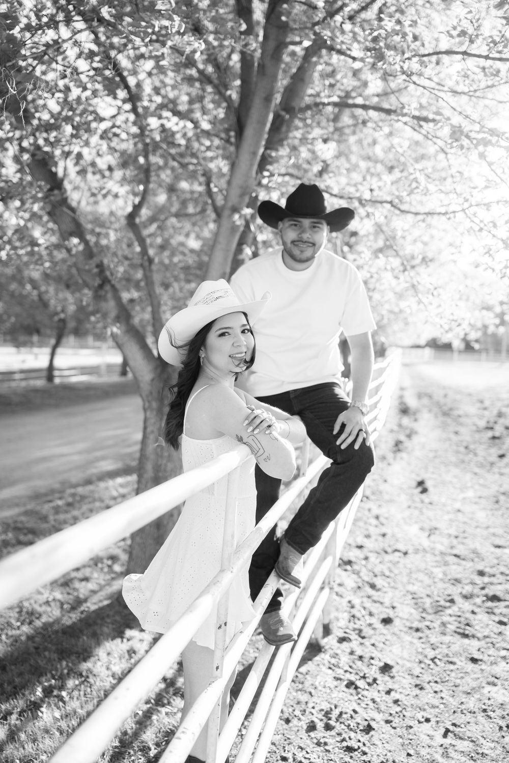 Black and white photo of a couple in cowboy hats posing by a white fence under blooming trees.