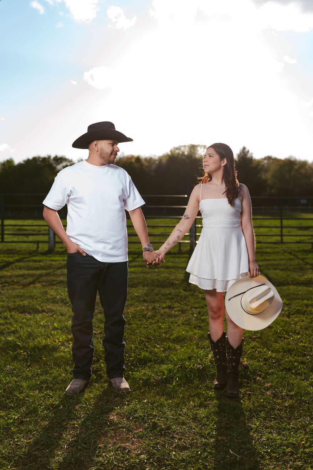 Couple holding hands in a green farm field, man in black cowboy hat and white tee, woman in white dress with boots.