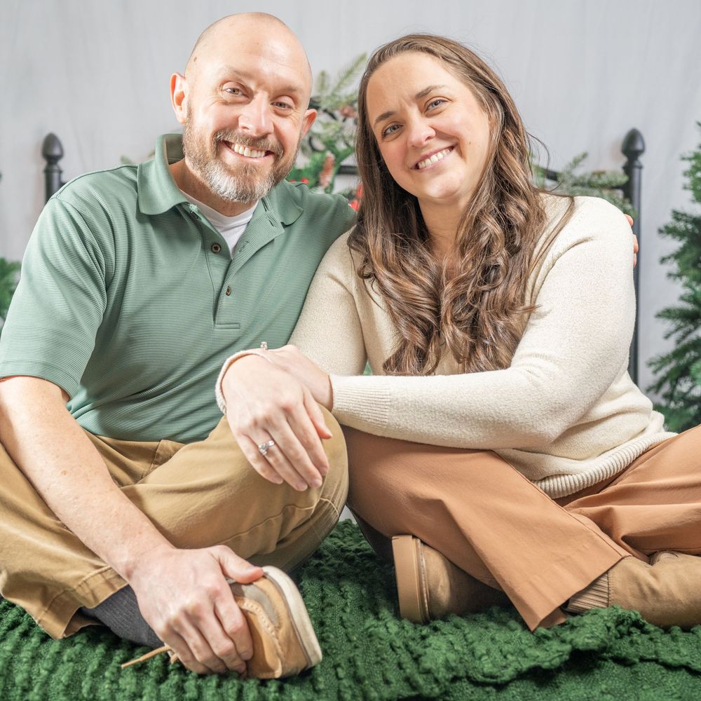 Smiling couple sitting together on green rug near Christmas tree in cozy holiday photo session.