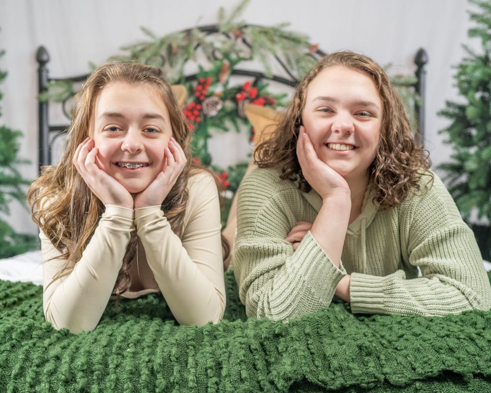 Two smiling teenage girls posing together on a bed with a festive Christmas backdrop and green blanket.