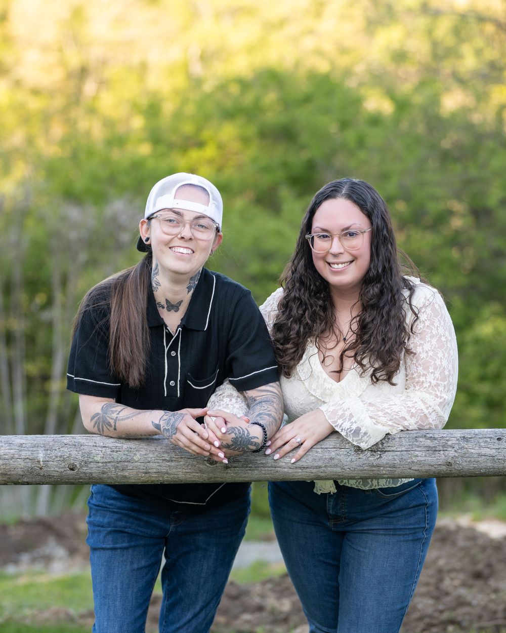 Two smiling women with tattoos lean on a wooden fence rail outdoors with green trees in the background.