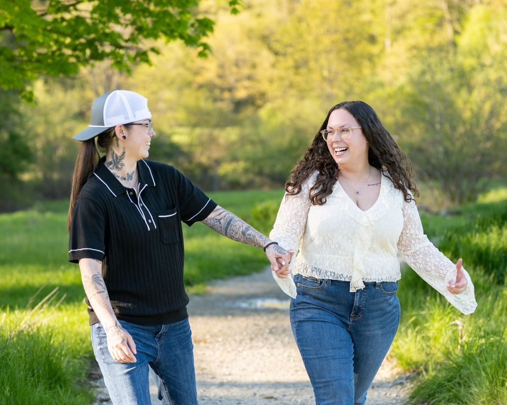 Two women holding hands and laughing while walking on a park path surrounded by lush green trees.