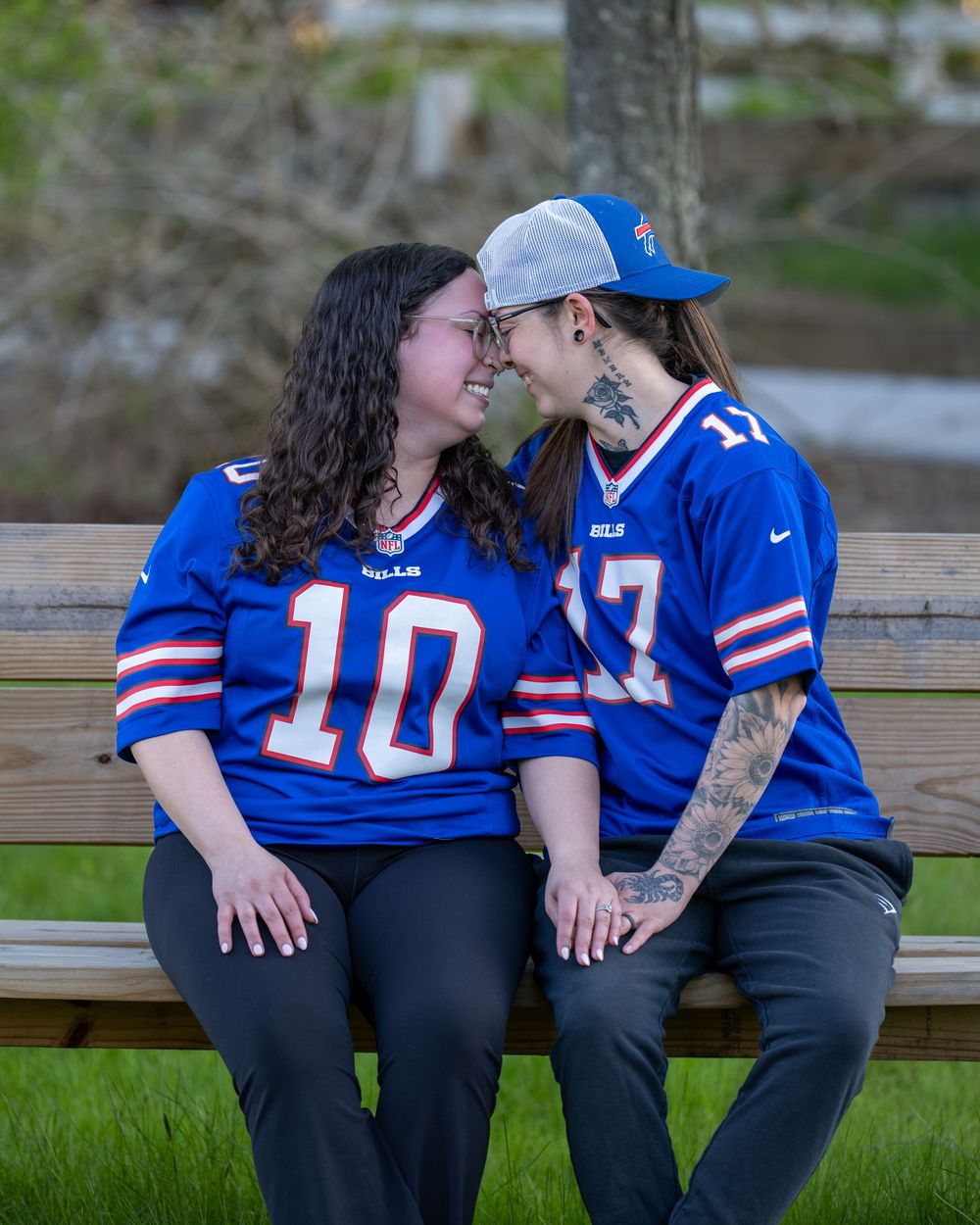 Two women in matching Buffalo Bills jerseys sit on a bench, smiling at each other in a romantic moment outdoors.
