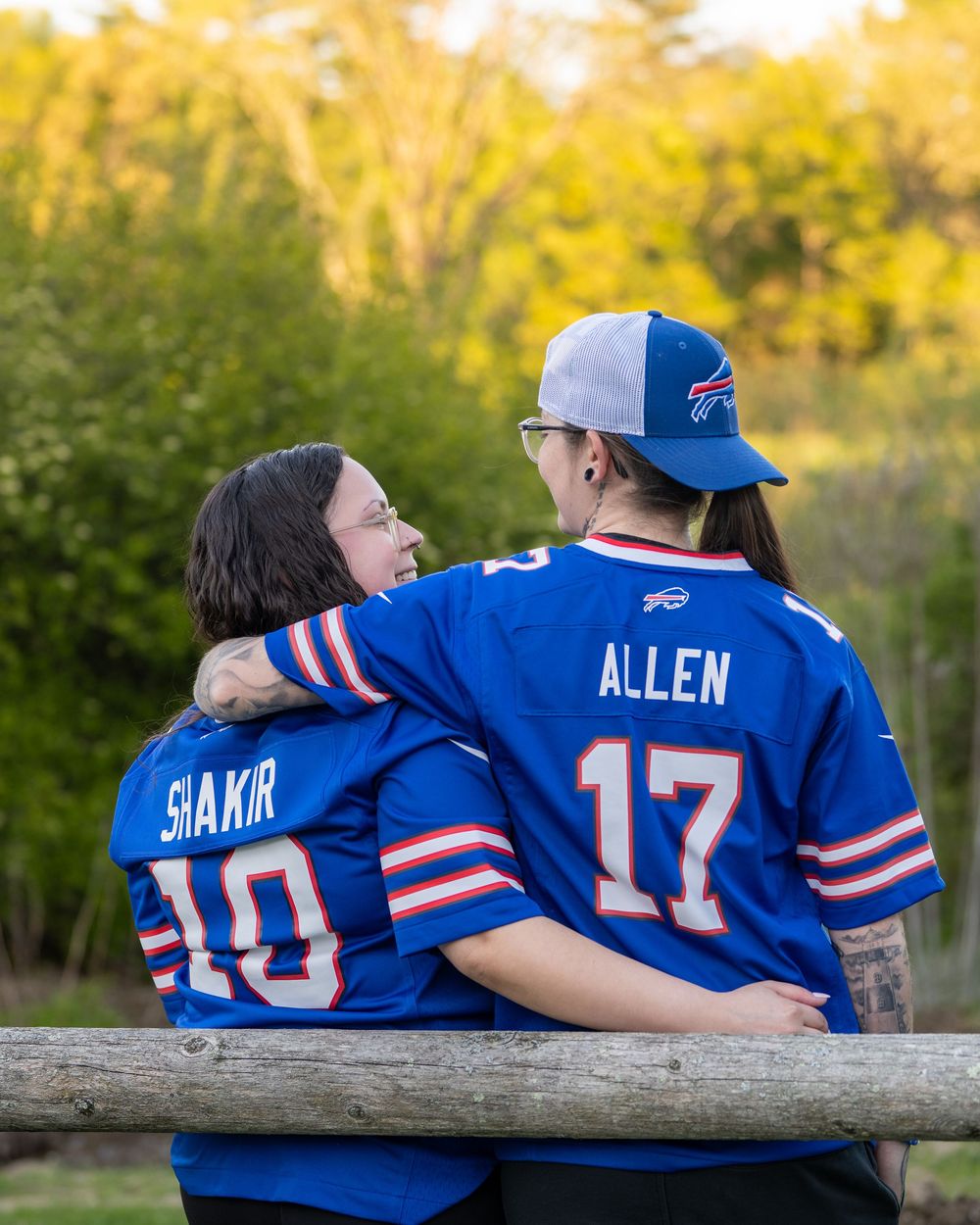 Two Buffalo Bills fans wearing Shakir #10 and Allen #17 jerseys sit on a log bench surrounded by autumn trees.