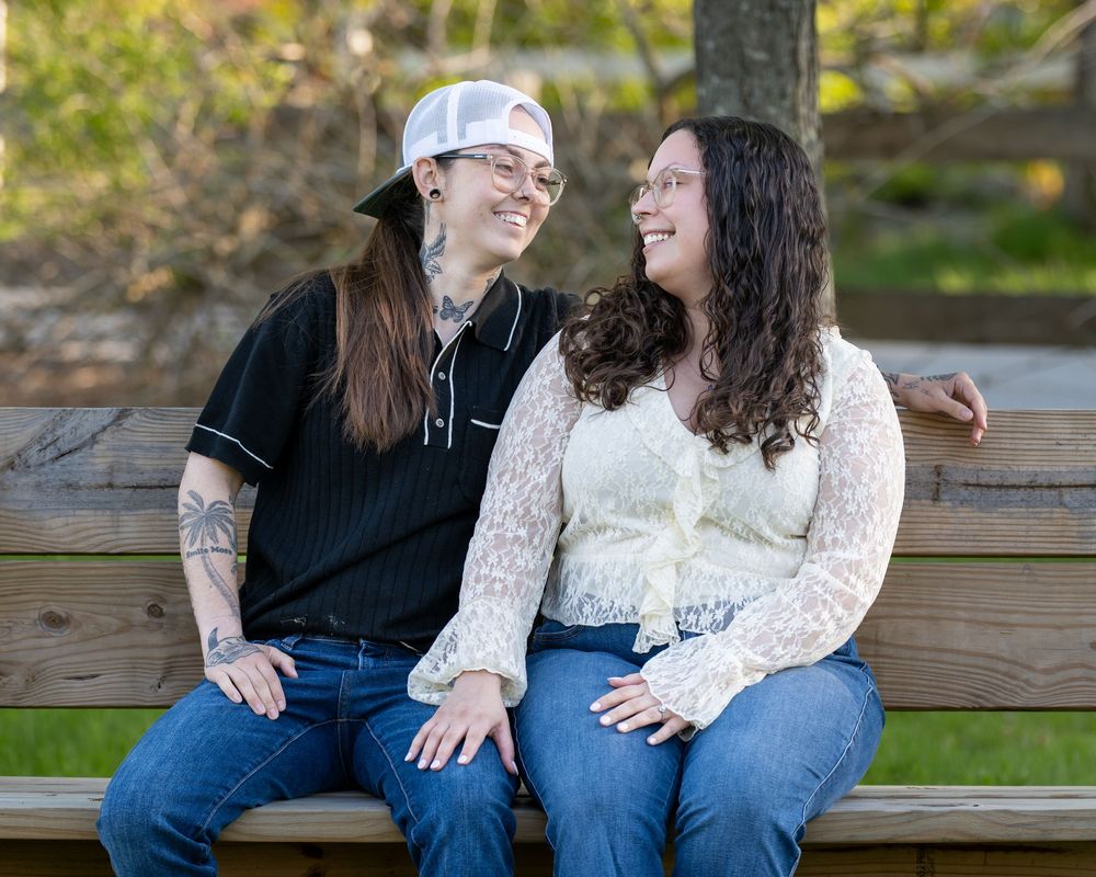 Two women sitting on a wooden bench outdoors, smiling at each other in a park setting.