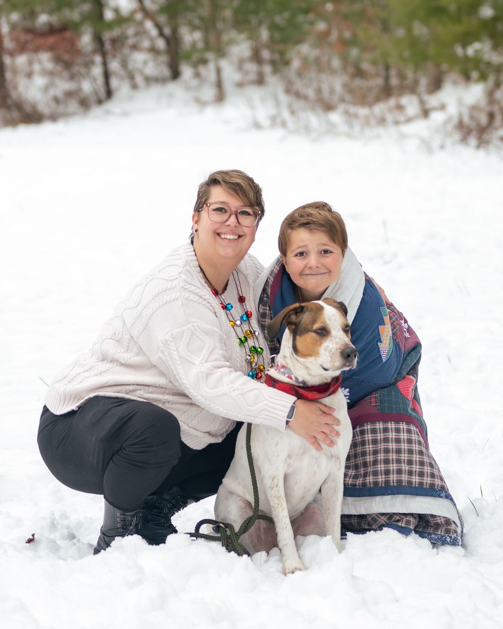 Two people and a dog pose together in snowy winter landscape, smiling warmly for a holiday family portrait.