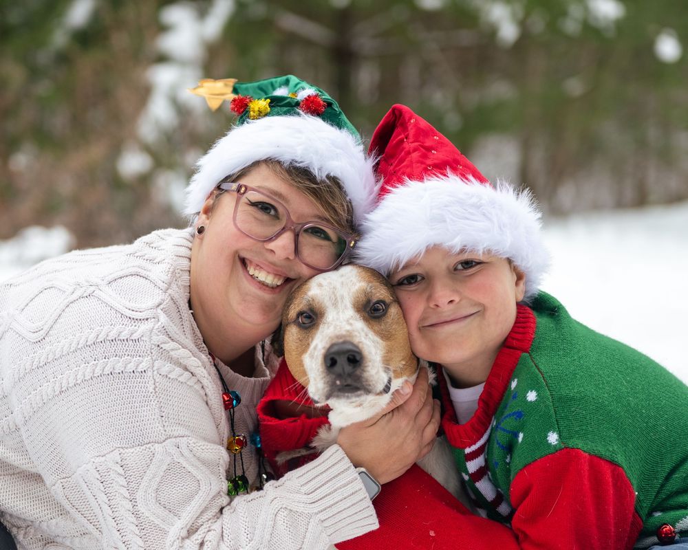 Two people in Christmas hats cuddle a beagle mix dog outdoors in a snowy winter setting.