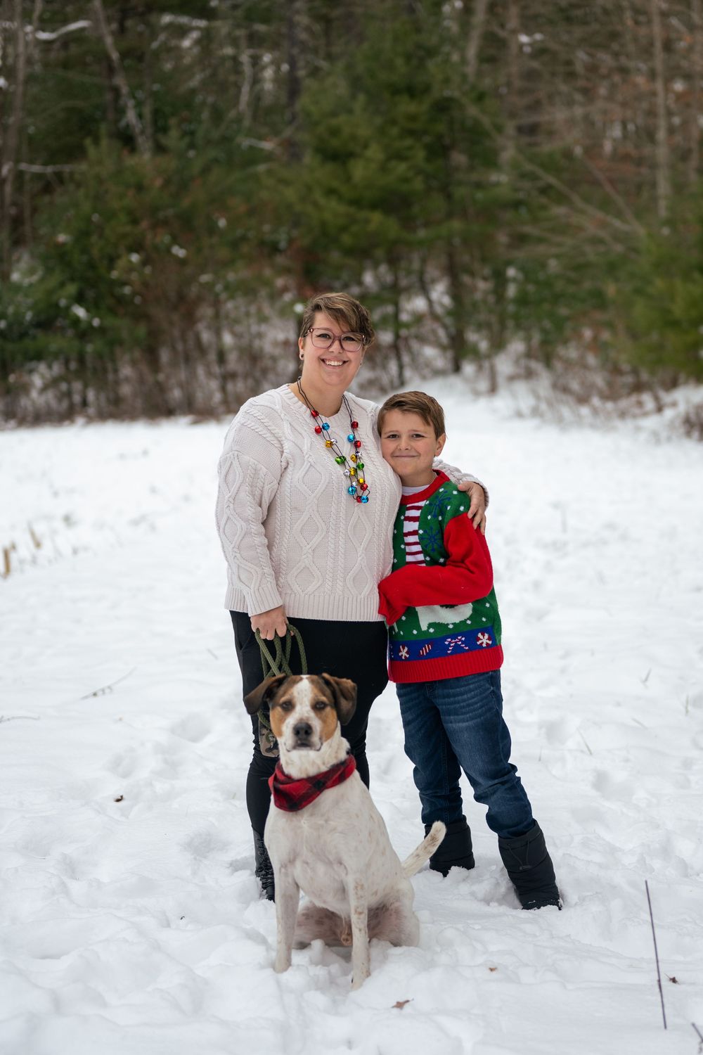 Woman and boy in holiday sweaters pose with their dog in a snowy winter forest setting.