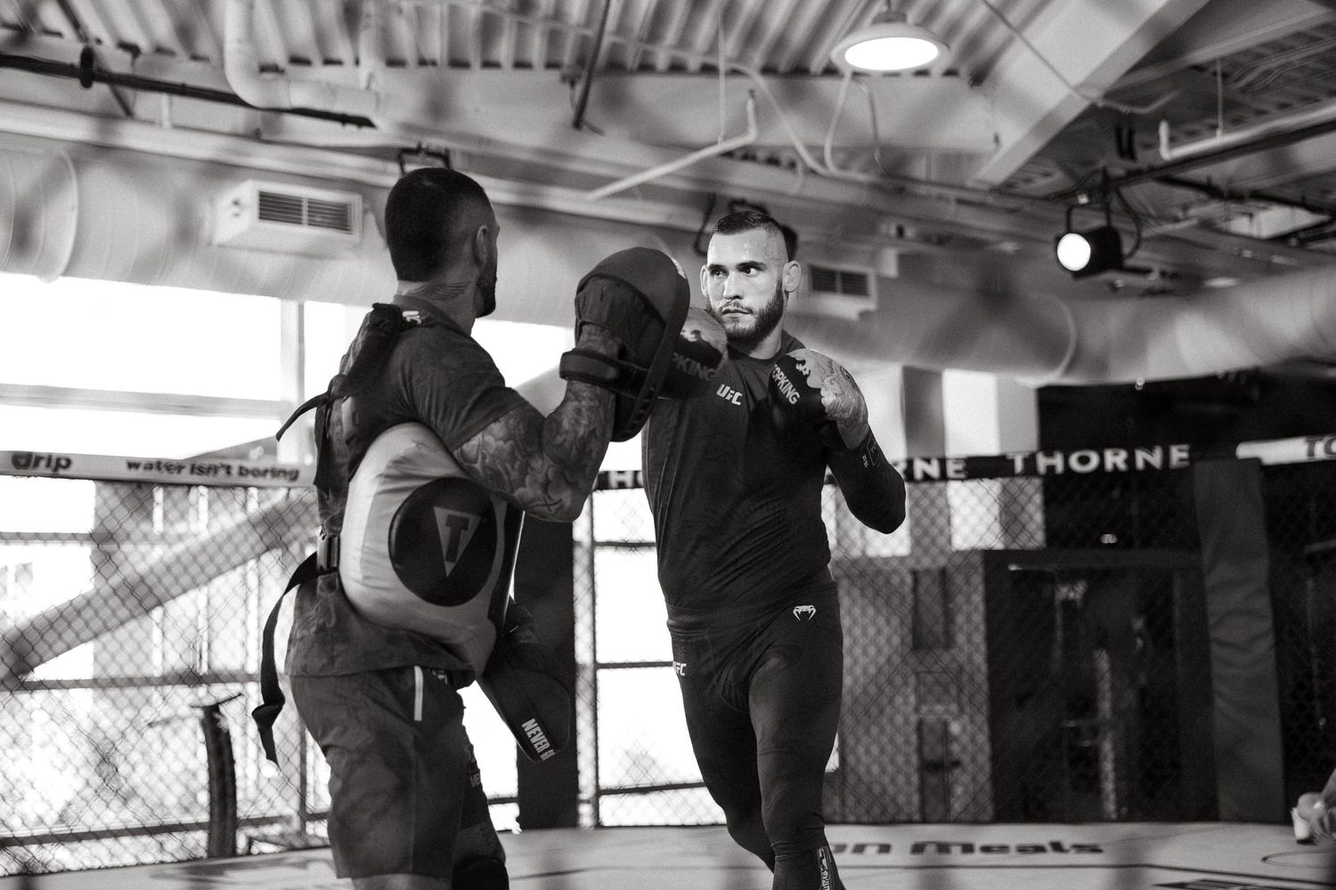 Two MMA fighters sparring in a gym, one throwing a punch while the other defends in a black and white photo.