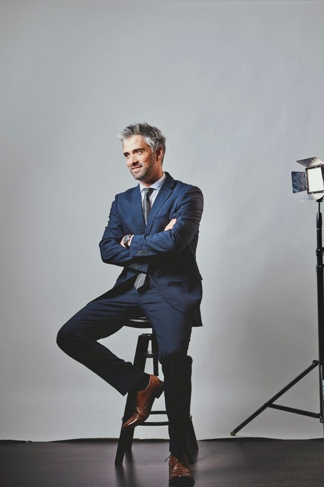 Confident businessman in a navy suit sitting on a stool with arms crossed in a professional studio.