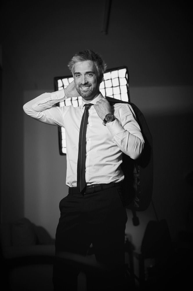 Smiling man in a white shirt and tie holding a jacket over his shoulder in a black and white photo.