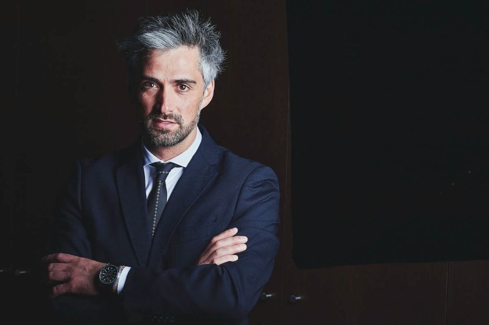 Serious businessman with gray hair in a navy suit sitting with arms crossed in a dimly lit setting.