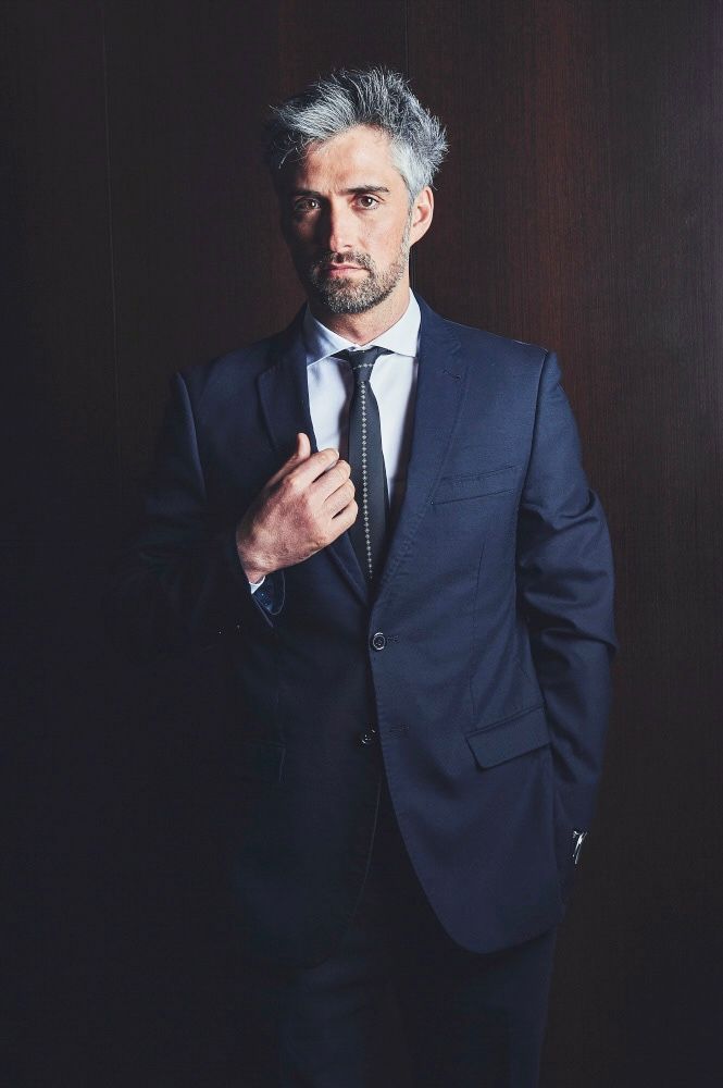 Confident businessman with gray hair in a navy suit adjusting his tie against a dark background.