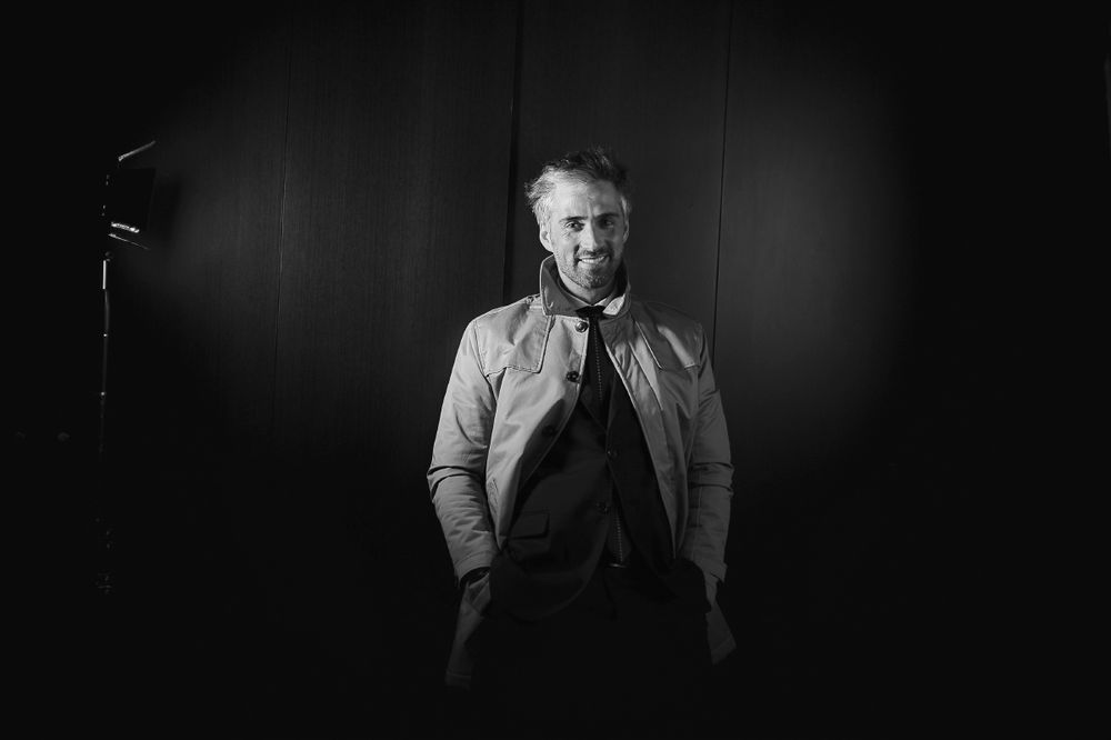 Black and white portrait of a mature man in a light jacket standing in dramatic studio lighting.