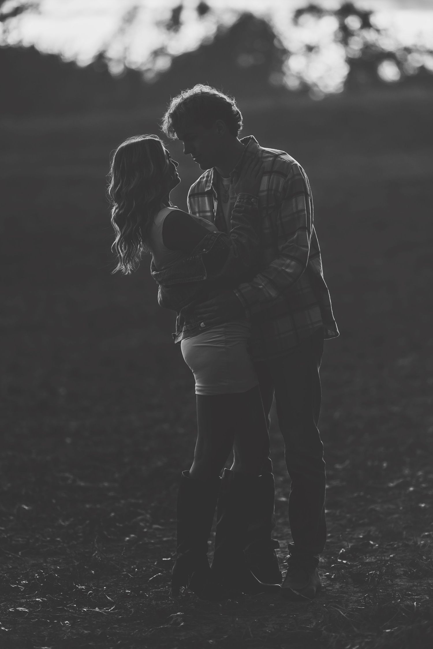 Black and white photo of a romantic couple embracing and kissing in an outdoor field at sunset.