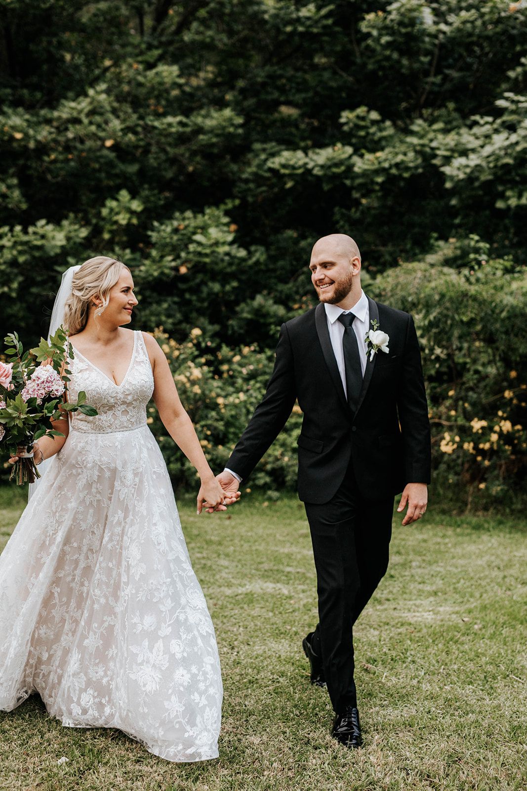 A smiling bride in a floral lace gown holds hands with a groom in a black suit as they walk through a lush garden.