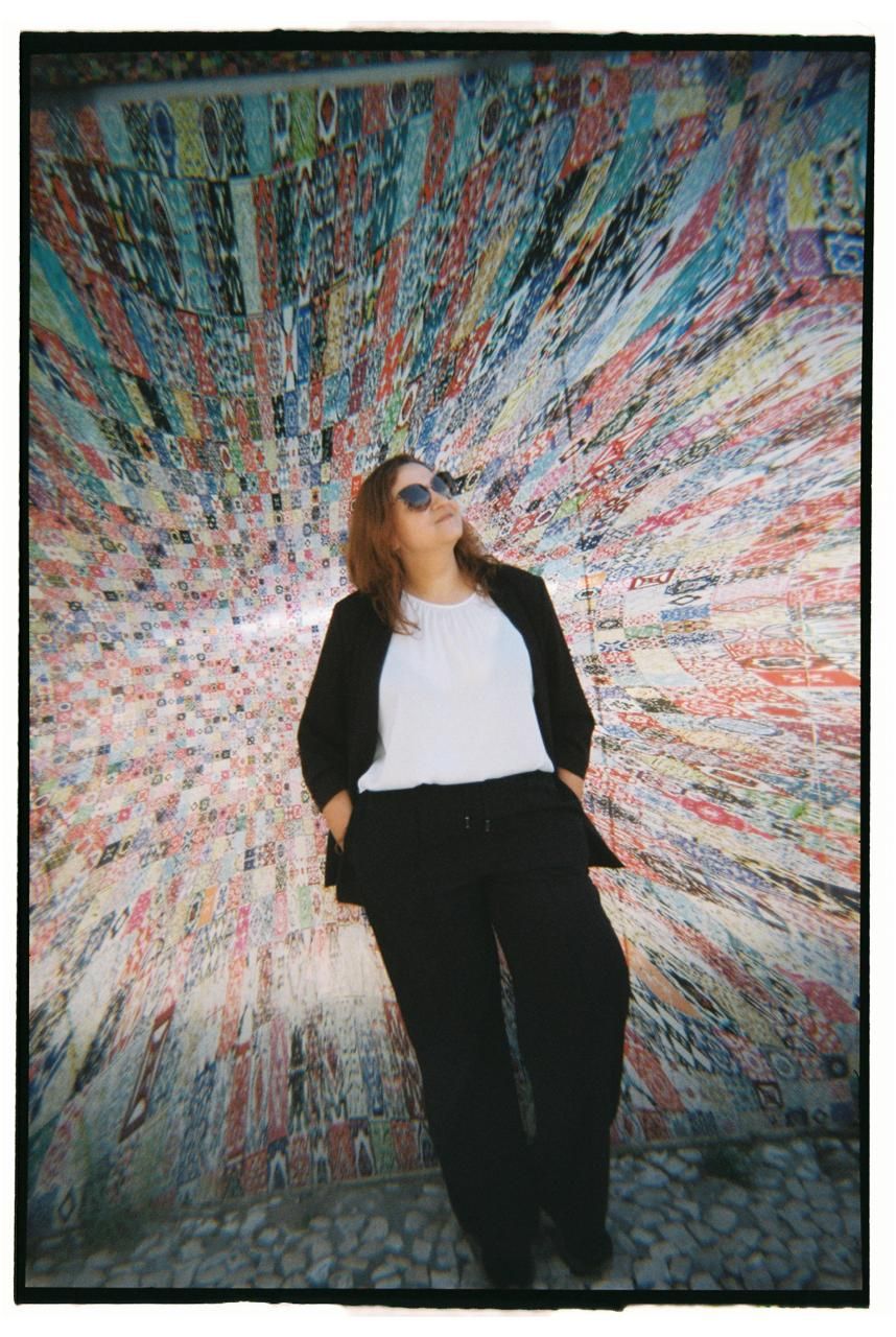 Woman in black outfit and sunglasses posing against a vibrant colorful mosaic wall art installation.