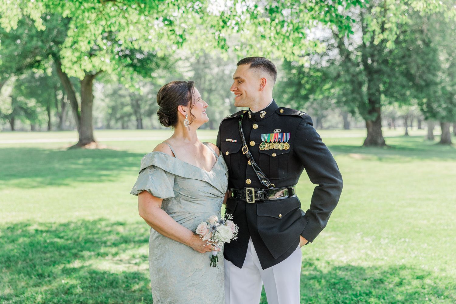 Military officer in dress uniform stands with elegant partner in gray gown outdoors in lush green park setting.
