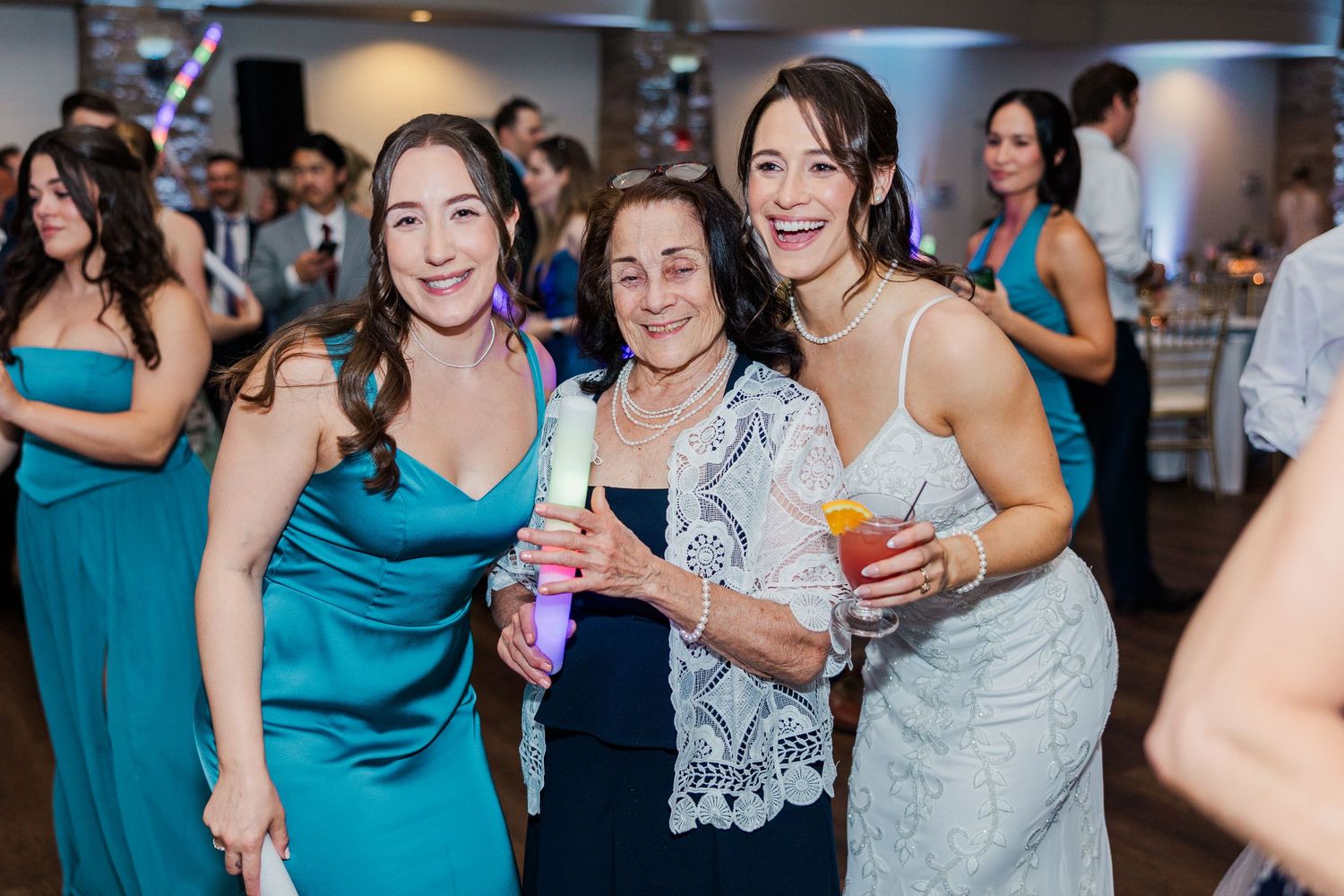 Three women smiling and posing together at a wedding reception, holding drinks and light-up accessories.