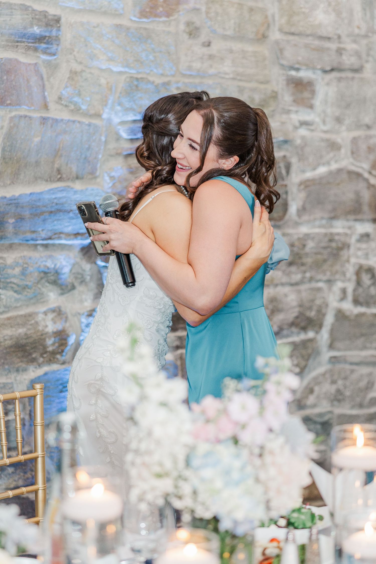 Bride in white lace gown hugging bridesmaid in teal dress at elegant wedding reception with candles and floral arrangements.