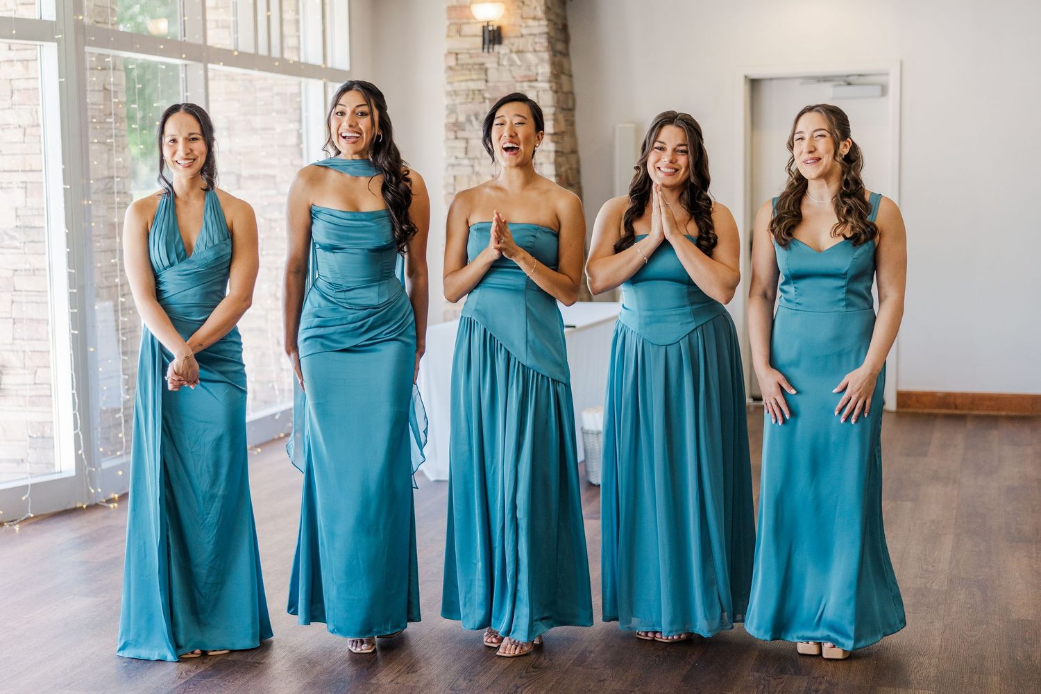 Five bridesmaids in matching teal satin gowns laughing and posing together in an elegant wedding venue.