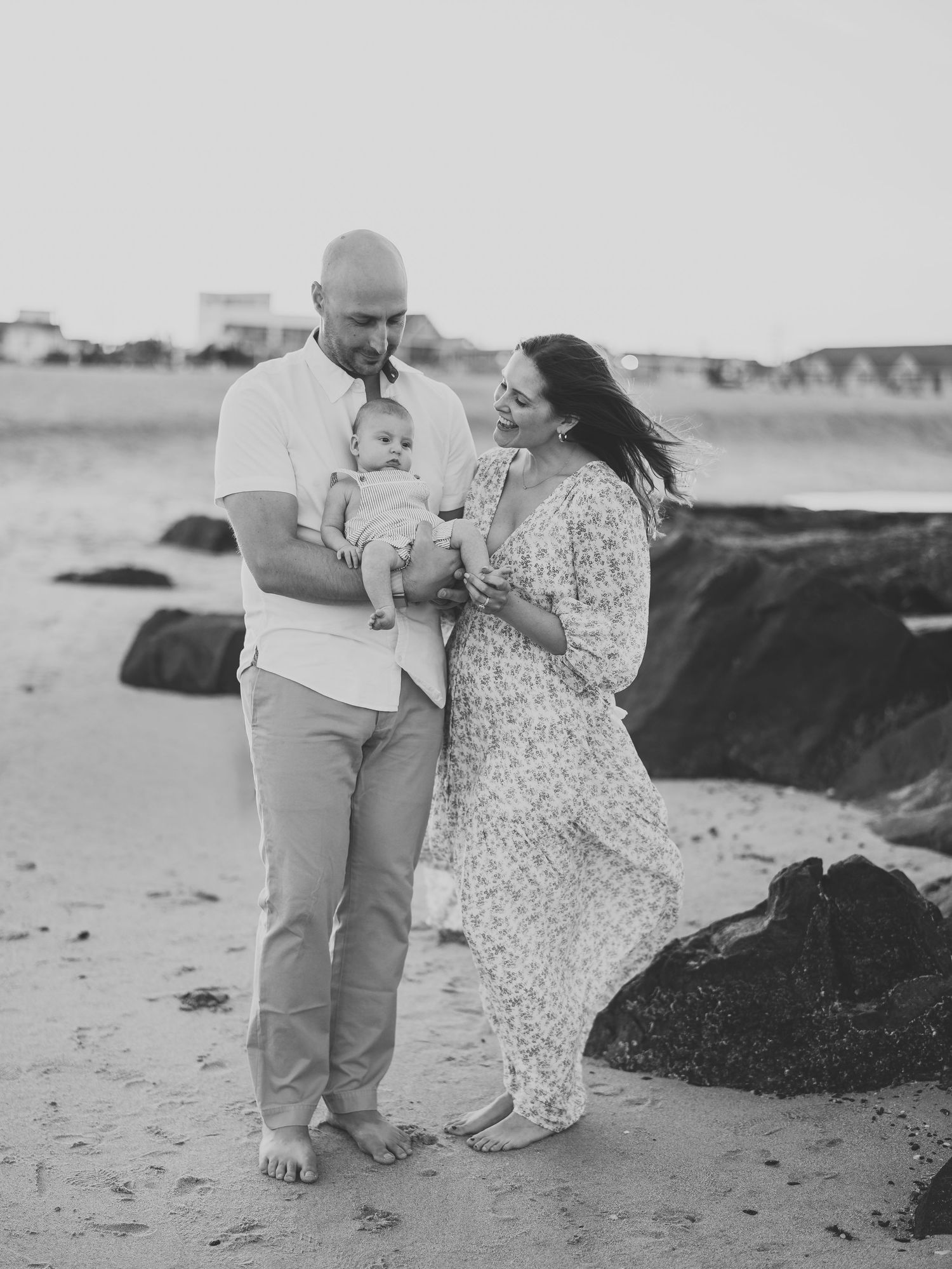 Belmar NJ newborn photographer capturing parents cuddling baby on the sand against a golden sunset sky in a black and white.
