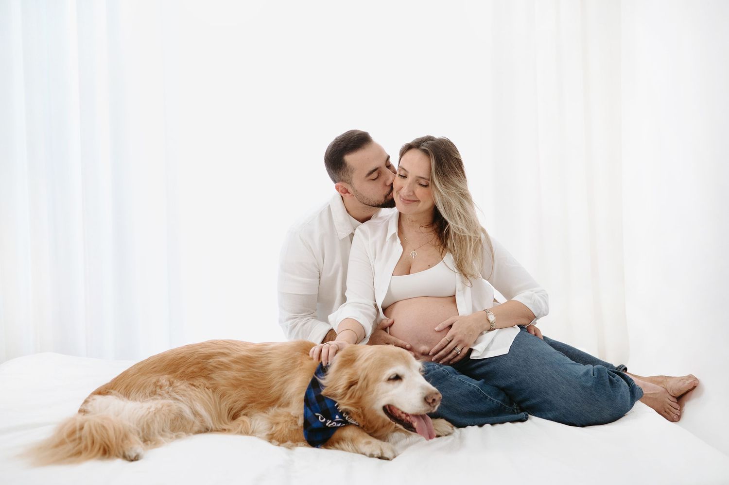 Pregnant couple sitting on white bed with golden retriever dog wearing blue bandana during maternity photo session.