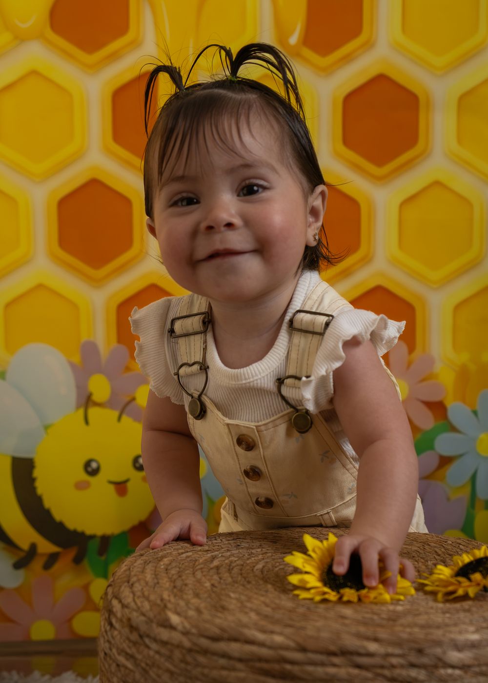 Smiling baby girl in overalls at a bumble bee themed first birthday party with honeycomb backdrop.