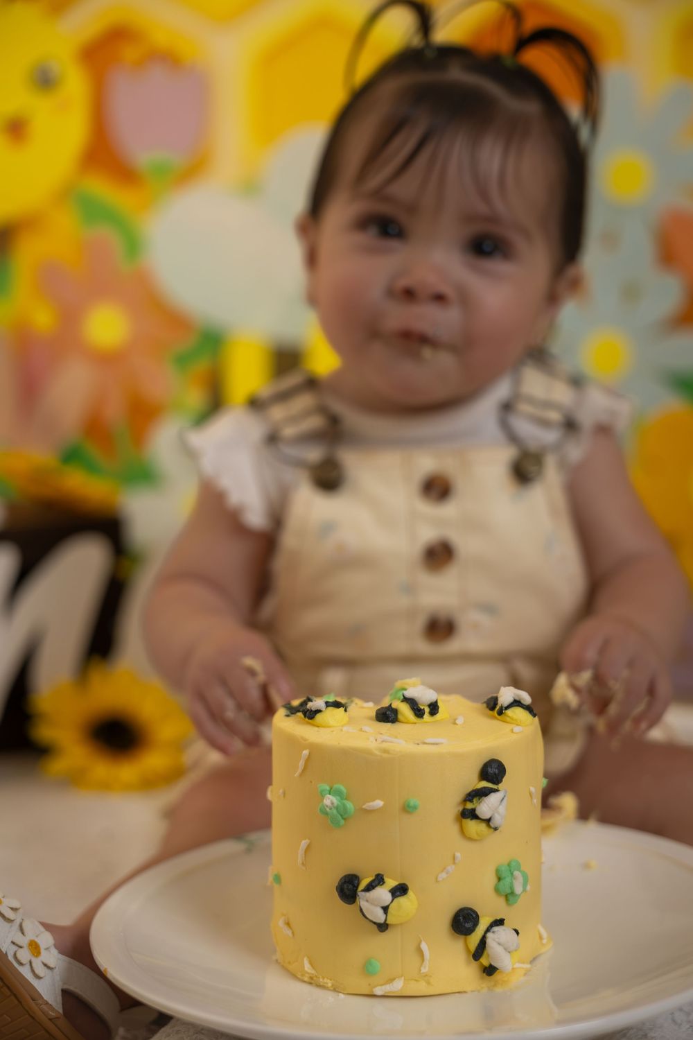 Baby girl in bee costume overalls with a yellow bumble bee smash cake on a white plate.