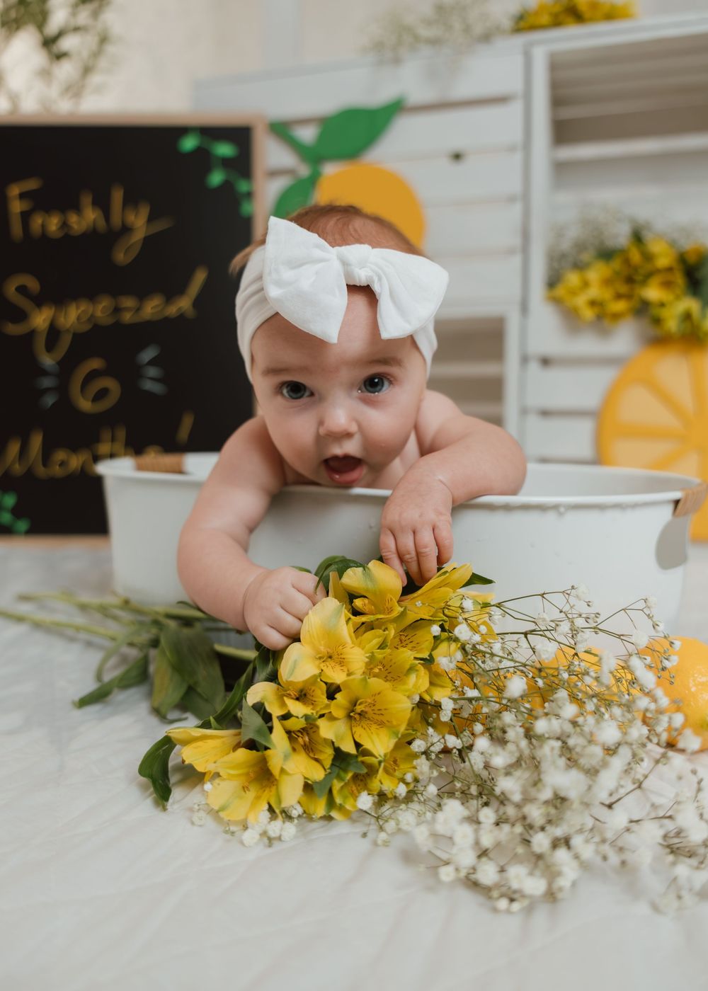 Six month old baby girl in white bow headband leaning over tub with yellow flowers indoors.