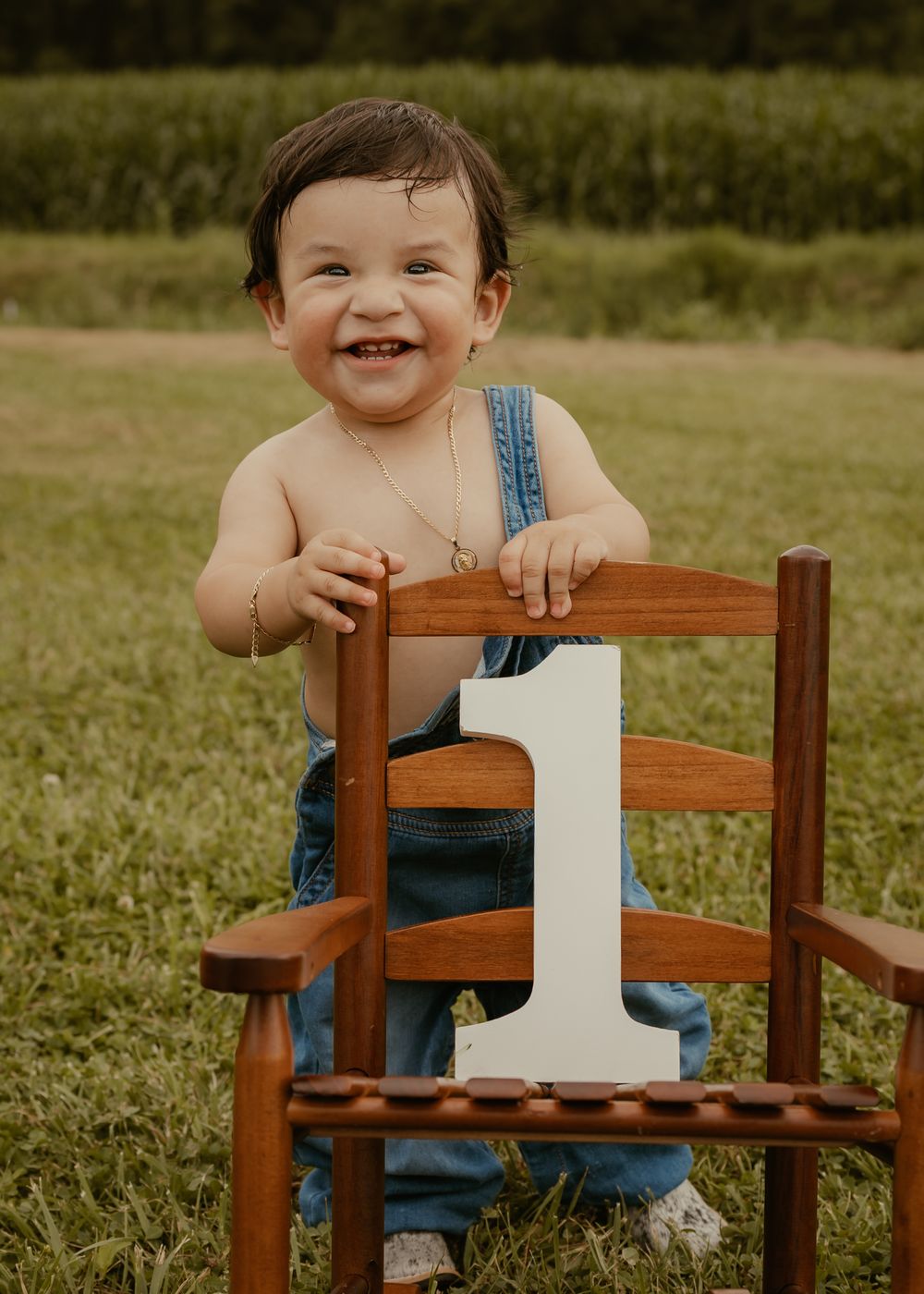 Smiling one year old boy in denim overalls standing at wooden chair holding number one sign.