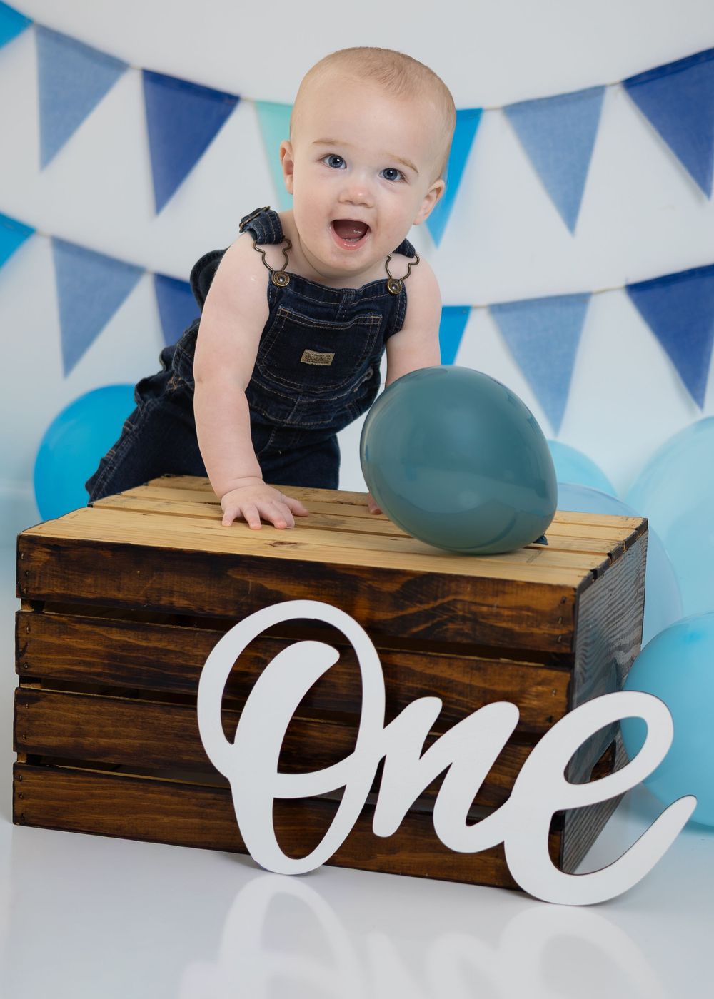Laughing baby in denim overalls stands at wooden crate with 'One' sign at blue-themed first birthday session.