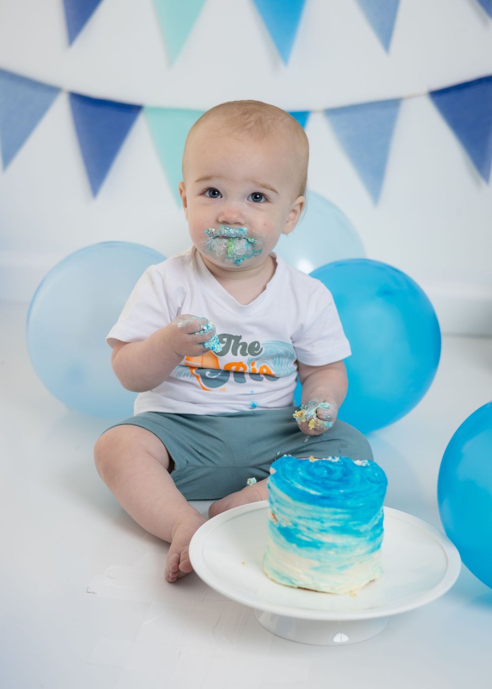 Baby with cake-covered face smashes blue ombre cake at a blue-themed first birthday cake smash photo session.