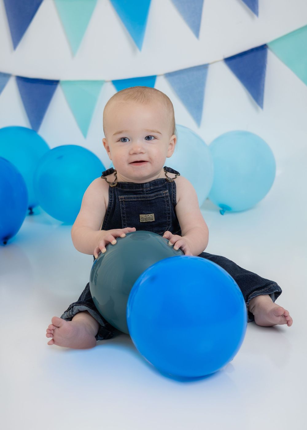 Baby in denim overalls plays with blue balloons at a blue-themed first birthday cake smash photo session.