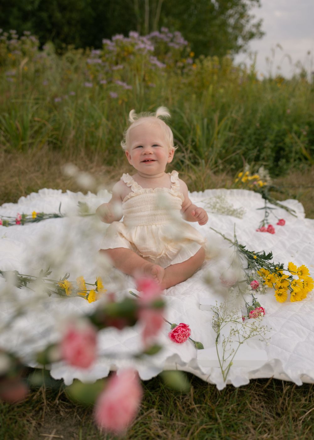 Happy baby girl in cream romper sitting on white blanket surrounded by wildflowers outdoors.