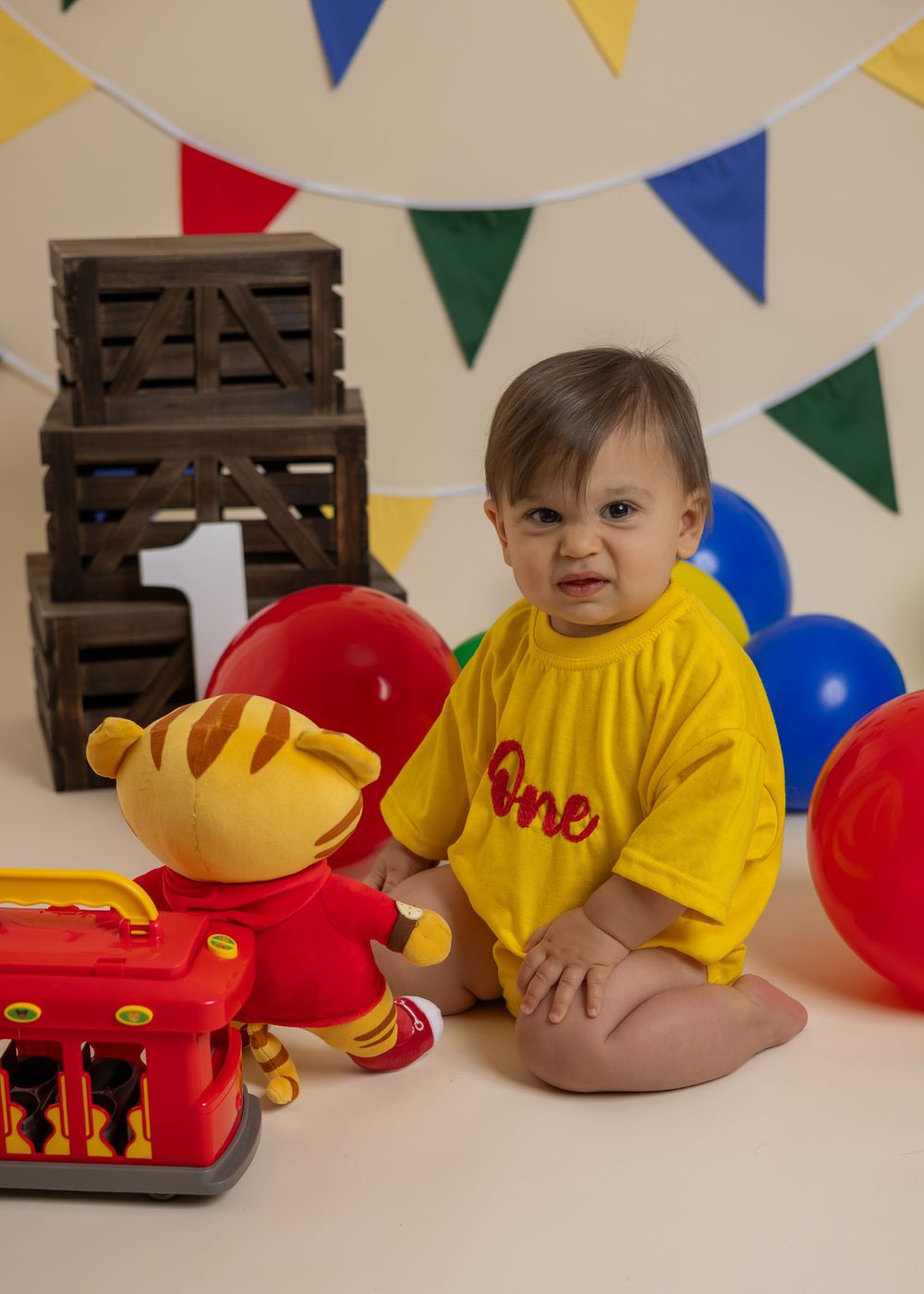 Baby in yellow 'One' onesie sits with Daniel Tiger toy and wooden crates at a first birthday cake smash photo session.
