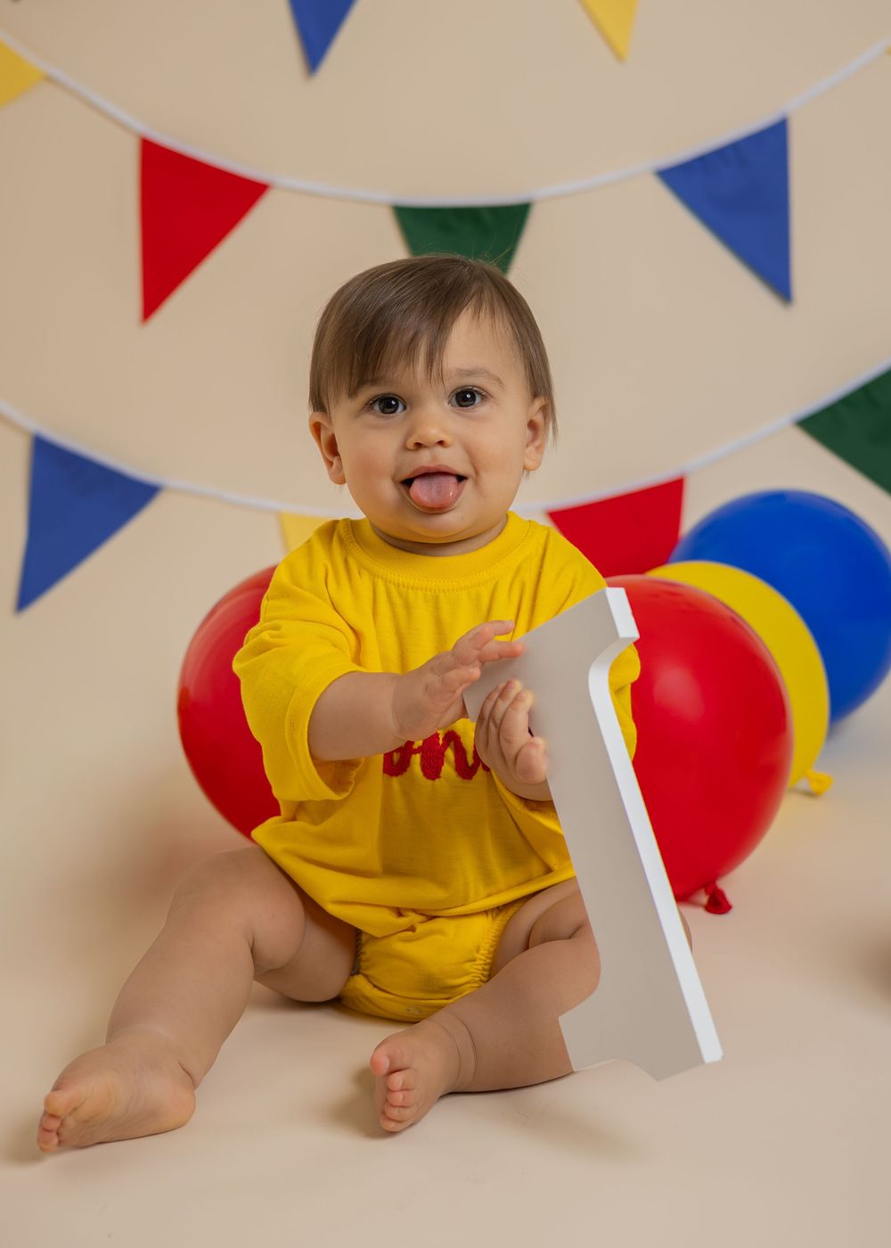 Smiling baby in yellow 'One' onesie reaches for number one sign at colorful first birthday photo session.
