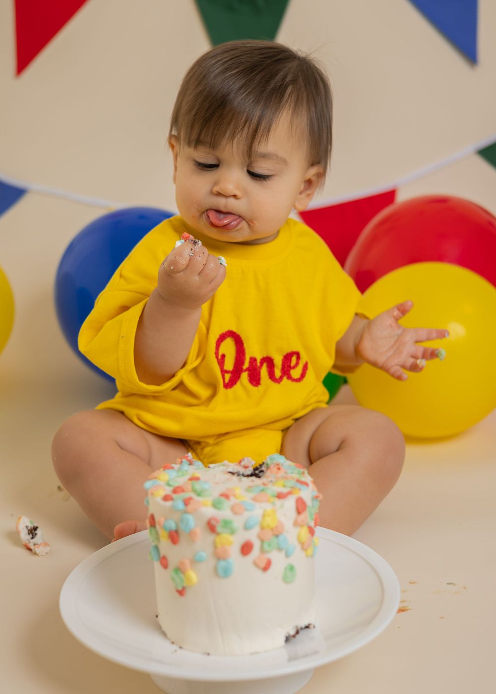 Baby in yellow 'One' onesie smashes colorful confetti cake at first birthday cake smash photo session.