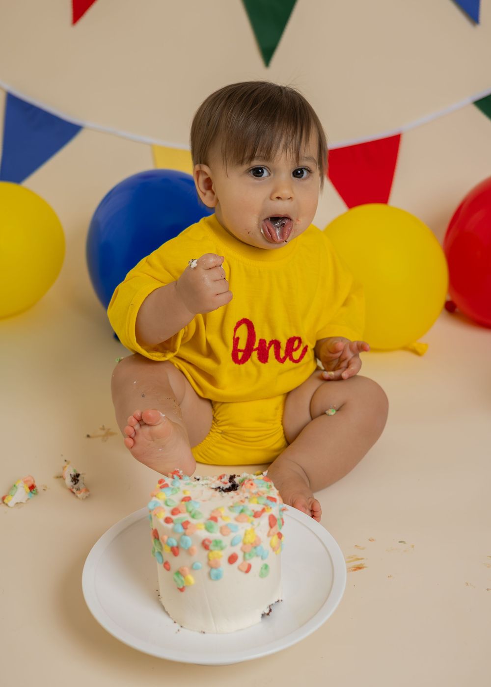 Baby in yellow 'One' onesie eats first birthday cake surrounded by colorful balloons and bunting flags.