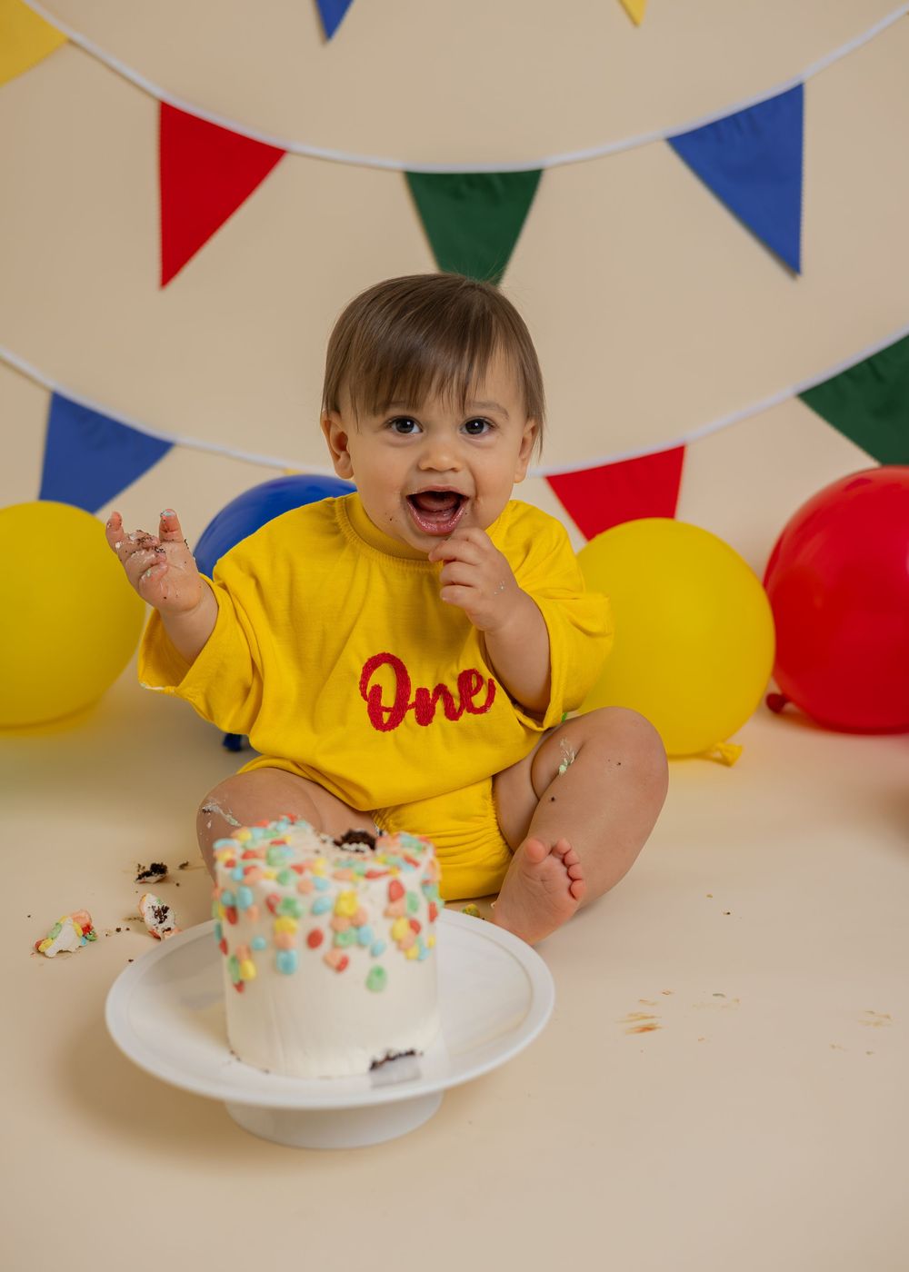 Happy baby in yellow 'One' onesie laughs while smashing colorful confetti cake at first birthday photo session.
