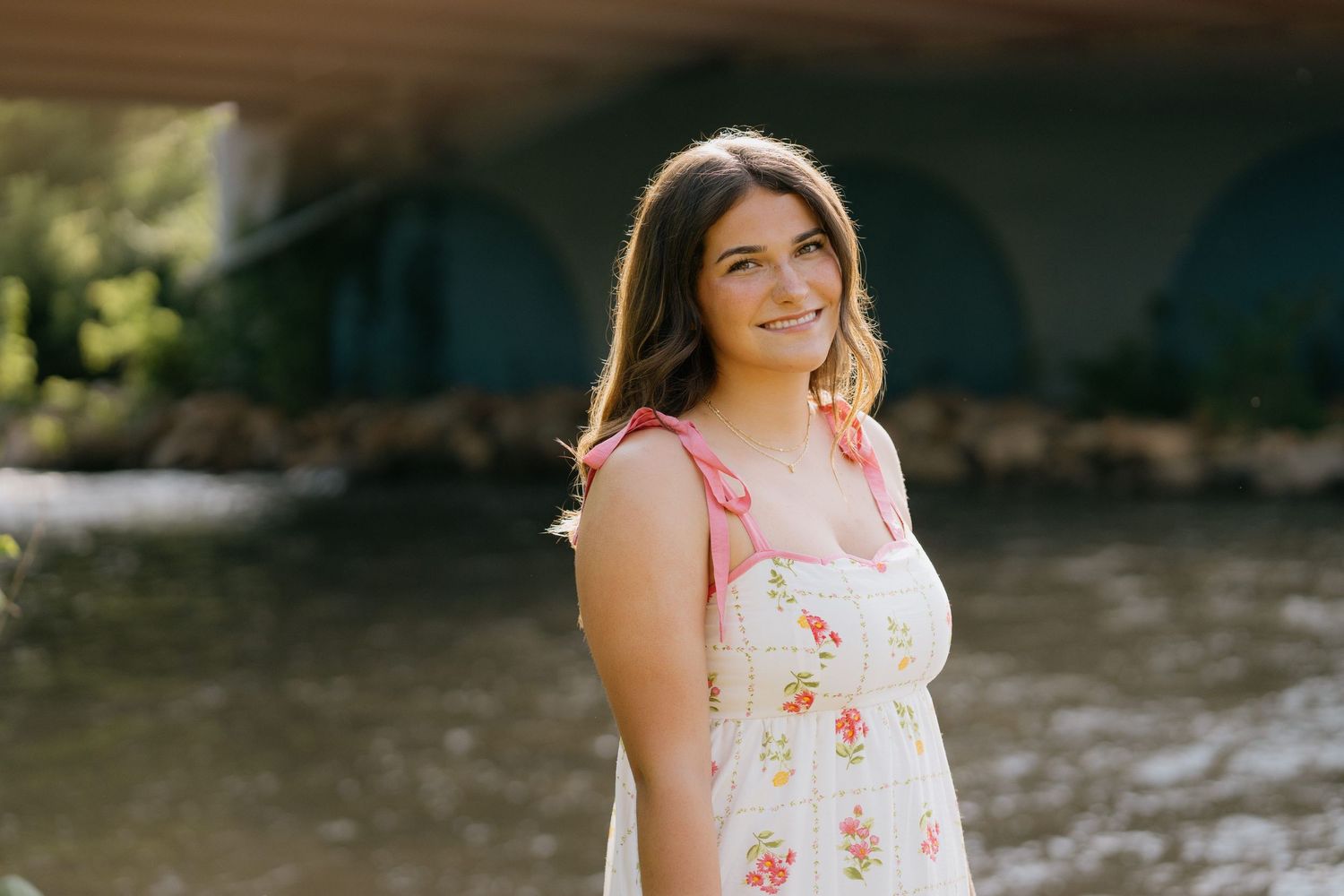 Young woman in floral sundress walking on a sunlit garden path surrounded by lush green trees.