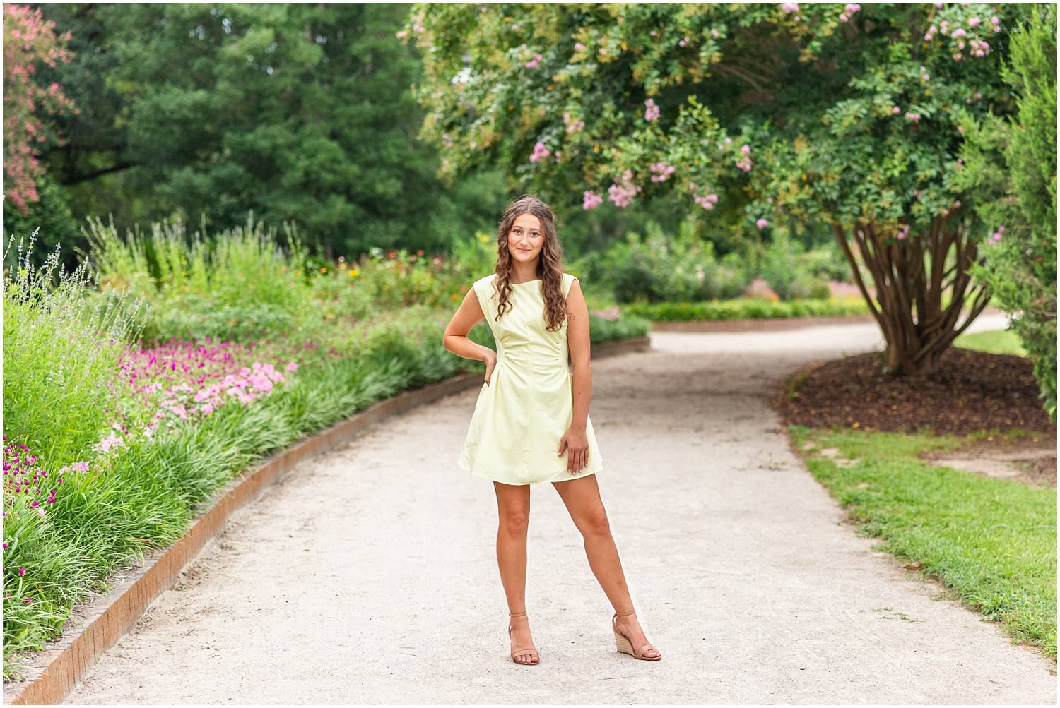 Young woman in light green mini dress poses on garden path surrounded by lush greenery and colorful flowers.
